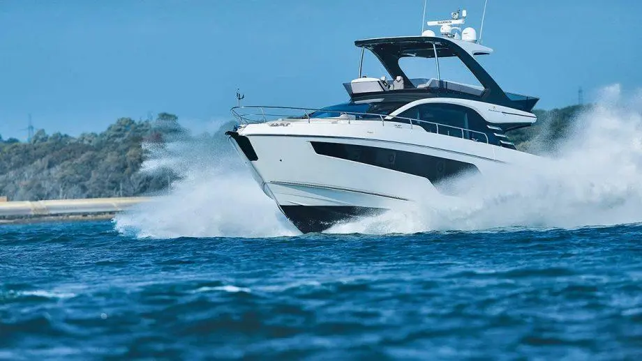 A sleek white luxury yacht glides through blue water, creating splashes against a backdrop of trees and clear sky.